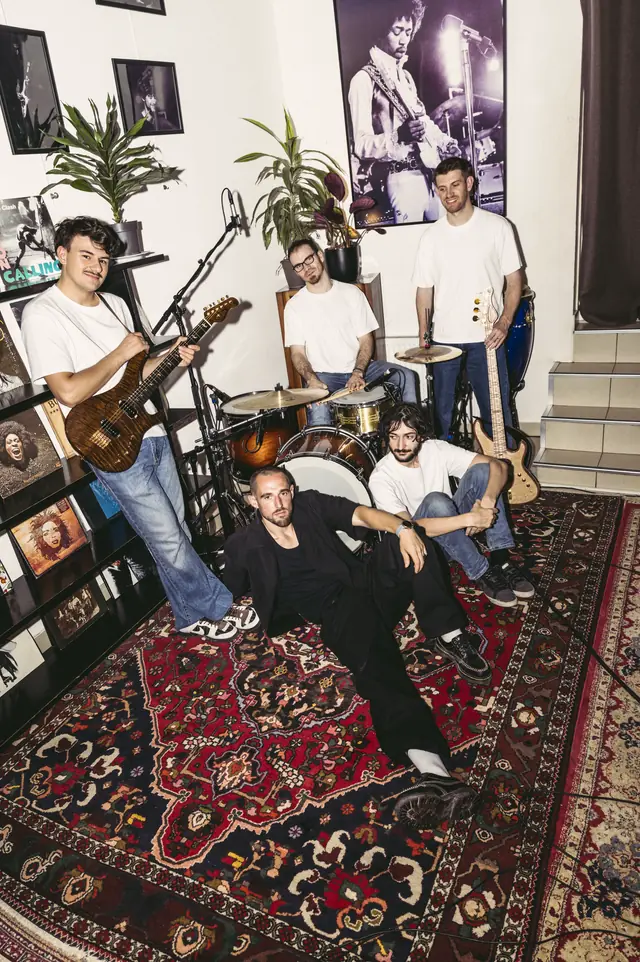 Les cinq membres du groupe posés dans le studio, guitare et basse en main, sur un grand tapis persan