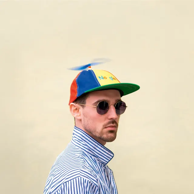Ex-Æquo de profil sur fond crème, lunettes rondes et casquette à hélice multicolore